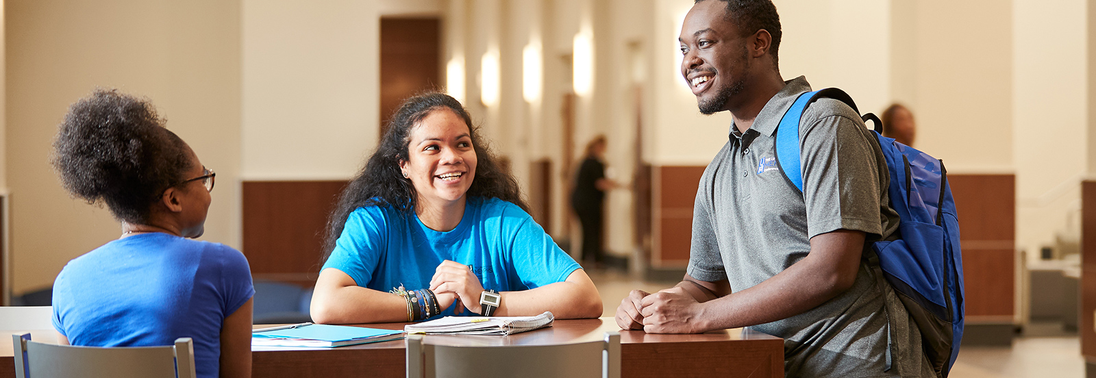 Three college students smiling and talking at a table in a bright campus lobby